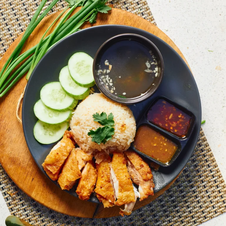Golden crispy fried chicken served with garlic rice, cucumber slices, hot soup, and two Thai-style dipping sauces on a dark plate