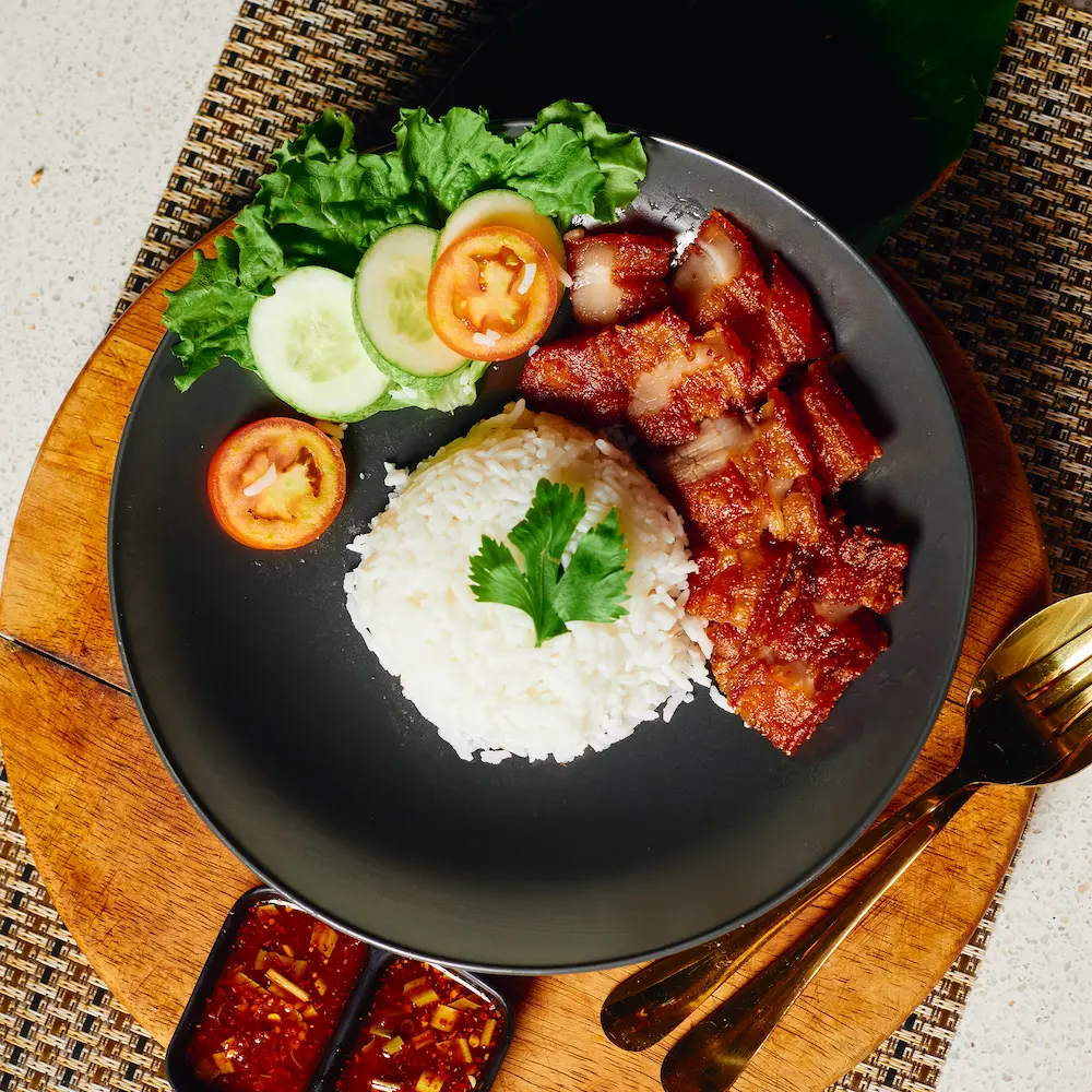 Crispy pork belly slices served with steamed jasmine rice, fresh cucumber, tomato slices, and spicy dipping sauces on a dark plate