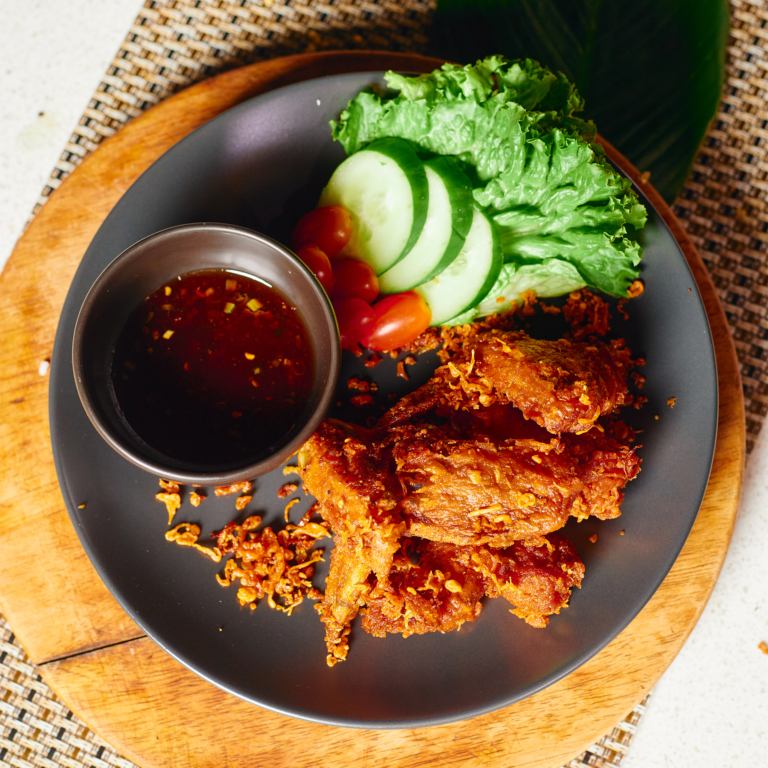 Thai-style fried chicken wings served with sweet chili sauce, cucumber, and fresh vegetables on a black plate.