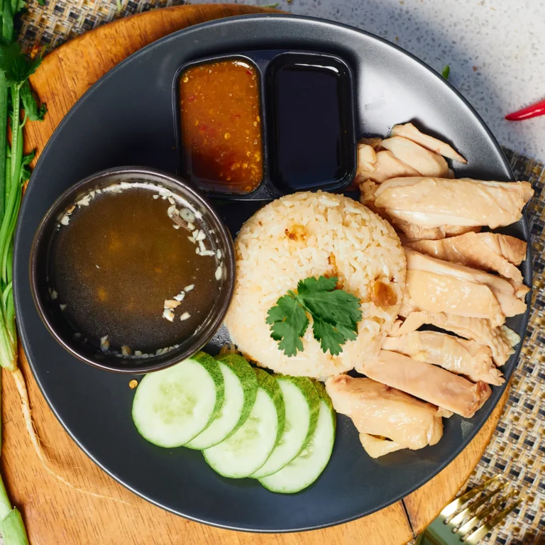 Steamed chicken served with fragrant rice, cucumber slices, savory soup, and two dipping sauces on a black plate