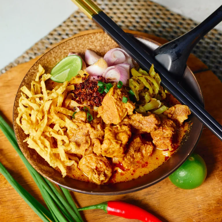 Northern Thai curry noodle soup with tender beef chunks, crispy noodles, shallots, lime, pickled mustard greens, and chili flakes