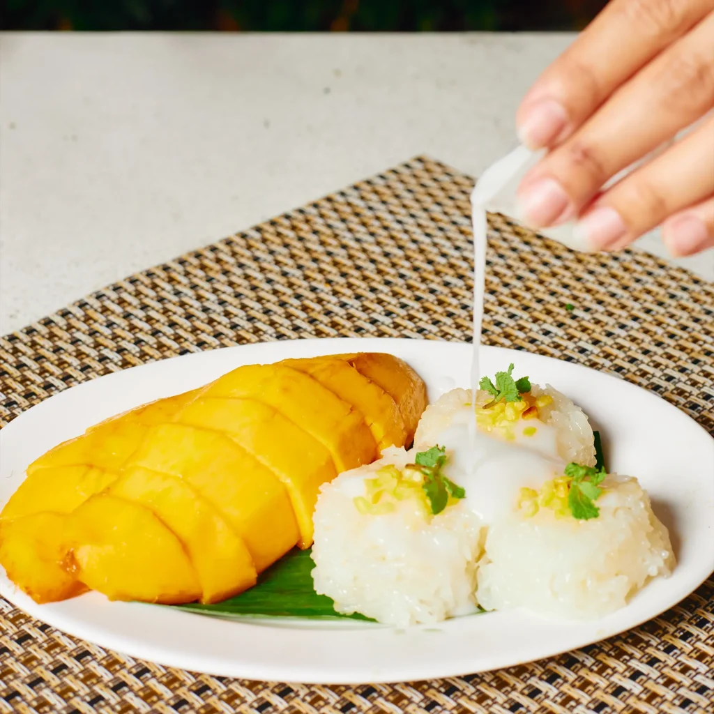 Traditional Thai mango sticky rice served with fresh ripe mango slices and coconut milk being poured over sweet glutinous rice