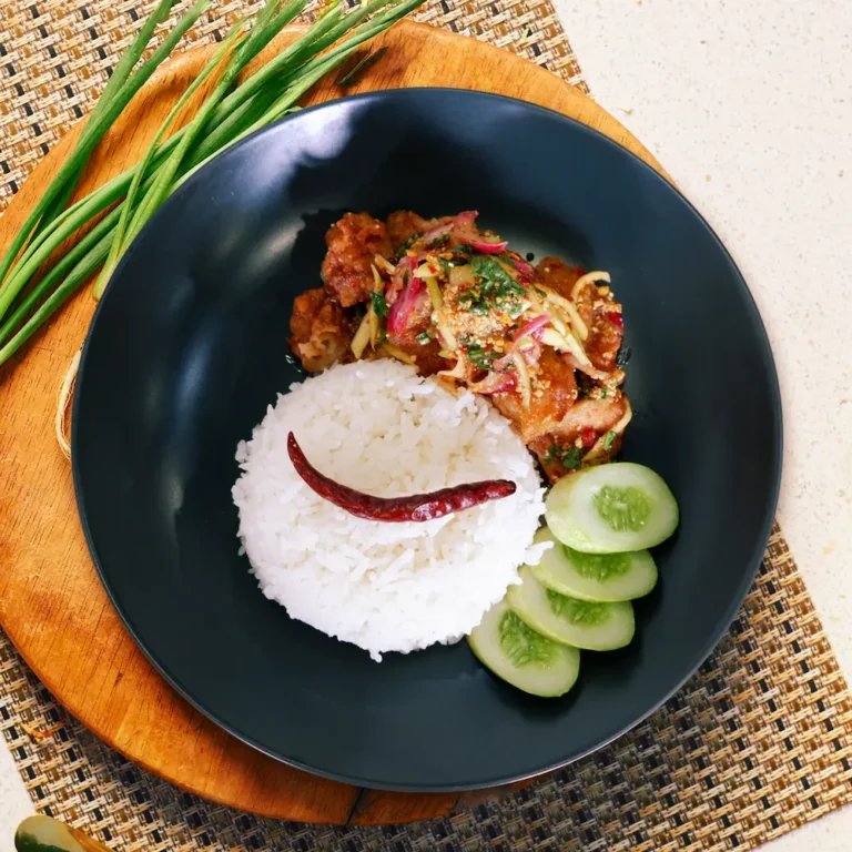 Spicy Thai-style chicken salad topped with herbs and chili, served with white jasmine rice and cucumber slices on a black plate